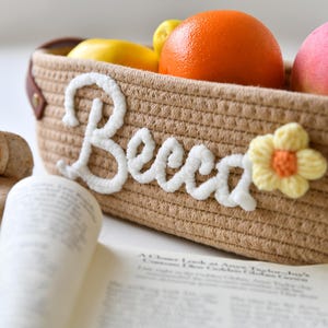 Op de afbeelding: Een bruine geweven mand met het woord "Boho" geborduurd in wit garen. De mand is gevuld met fruit, waaronder sinaasappels, citroenen en een roze perzik.