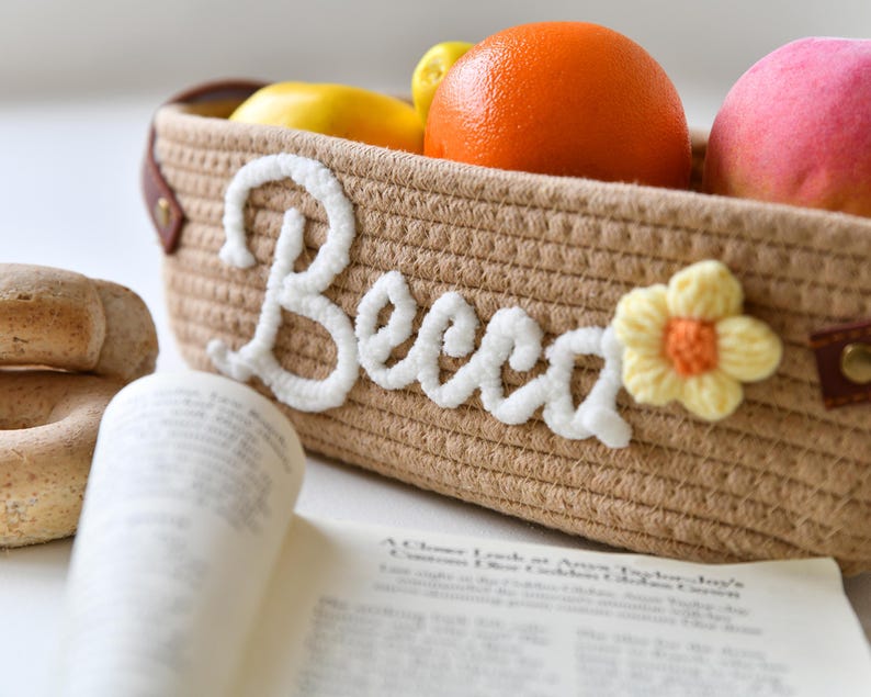 Op de afbeelding: Een bruine geweven mand met het woord "Boho" geborduurd in wit garen. De mand is gevuld met fruit, waaronder sinaasappels, citroenen en een roze perzik.