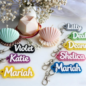 May include: A collection of personalized keychains in various colors, each with a name in white lettering. The keychains are attached to silver clasps. The image also features seashell-shaped candles and a geometric candle.