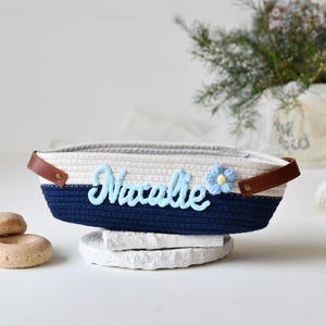 Op de afbeelding: Een witte en blauwe touwenmand met een bruin leren handvat. De mand heeft de naam "Natalie" geborduurd in blauw garen met een blauwe bloem.