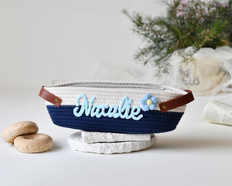 Op de afbeelding: Een witte en blauwe touwenmand met een bruin leren handvat. De mand heeft de naam "Natalie" geborduurd in blauw garen met een blauwe bloem.