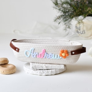 Op de afbeelding: Een witte touwenmand met bruine leren handvatten. De mand is geborduurd met de naam "Andrew" in pastelkleuren met een kleine oranje bloem.