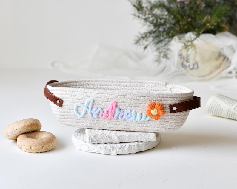 Op de afbeelding: Een witte touwenmand met bruine leren handvatten. De mand is geborduurd met de naam "Andrew" in pastelkleuren met een kleine oranje bloem.