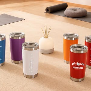 May include: A collection of insulated tumblers in various colors, including blue, purple, white, orange, and red. Each tumbler has a silver lid and base. Some tumblers feature personalized names or designs in a cursive font. The tumblers are designed for beverages.