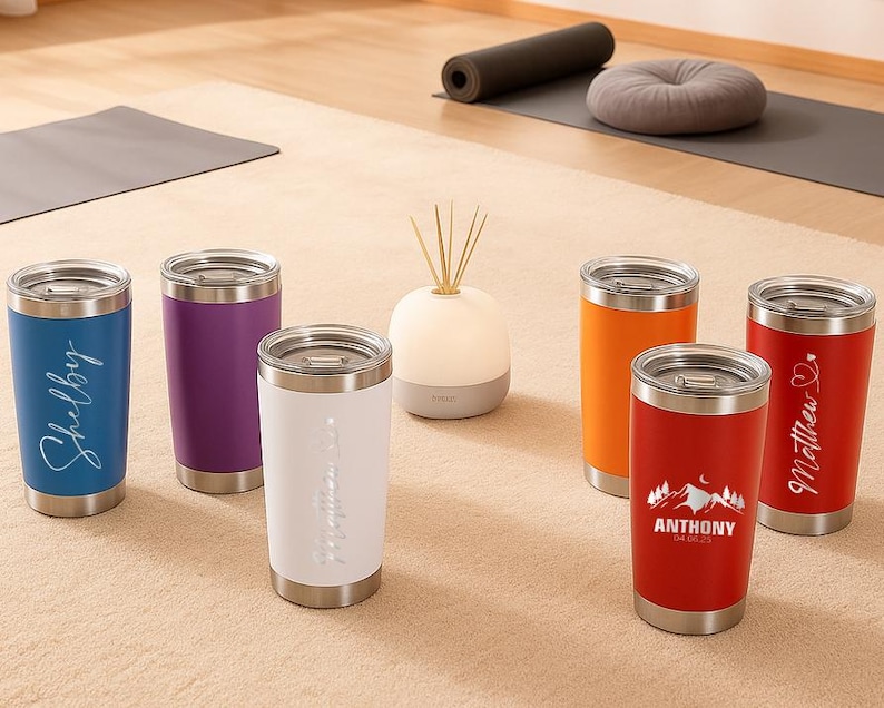 May include: A collection of insulated tumblers in various colors, including blue, purple, white, orange, and red. Each tumbler has a silver lid and base. Some tumblers feature personalized names or designs in a cursive font. The tumblers are designed for beverages.