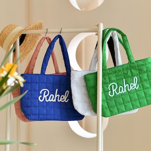 以下が含まれることがあります： 白で刺繍された「Rachel」という名前がついた、キルティングされたトートバッグが3つ。バッグはラックに掛けられており、青、緑、白です。
