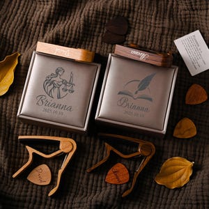 Könnte beinhalten: Zwei personalisierte Geschenkboxen mit Holzdeckeln, graviert mit Namen und Daten, sind zu sehen. Jede Box hat ein anderes Design, eine mit einer Waage und die andere mit einer Feder und einem Buch. Gitarrenkapodaster und Plektren sind ebenfalls sichtbar.