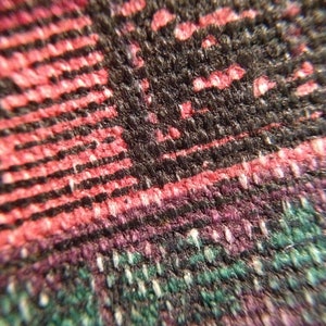 May include: Close-up of a woven fabric with a complex pattern. The fabric features a mix of colors, including black, red, purple, and green. The texture appears to be coarse, with visible threads and a slightly raised surface. The pattern is a mix of stripes and other shapes.