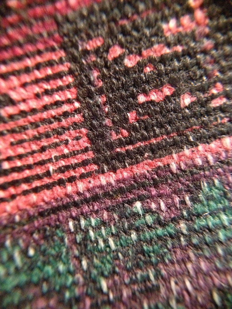 May include: Close-up of a woven fabric with a complex pattern. The fabric features a mix of colors, including black, red, purple, and green. The texture appears to be coarse, with visible threads and a slightly raised surface. The pattern is a mix of stripes and other shapes.