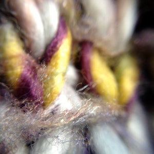 May include: Close-up of woven fabric with a mix of colors. The image highlights strands of yarn in yellow, purple, and white. The texture appears soft and fuzzy, with a focus on the intricate details of the weave.