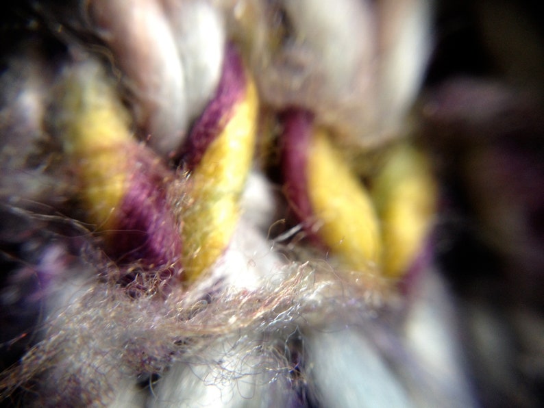 May include: Close-up of woven fabric with a mix of colors. The image highlights strands of yarn in yellow, purple, and white. The texture appears soft and fuzzy, with a focus on the intricate details of the weave.