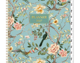 May include: A spiral-bound 2027 planner with a light blue cover featuring a pattern of pink flowers, green leaves, and various birds perched on brown branches. A central rectangular label displays the text PLANNER 2027 in gold lettering.