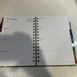 May include: An open spiral-bound planner showing the week of April 5 to April 11, 2027. The left page features a priorities list and days Monday through Wednesday. The right page lists Thursday through Sunday with lined writing spaces.
