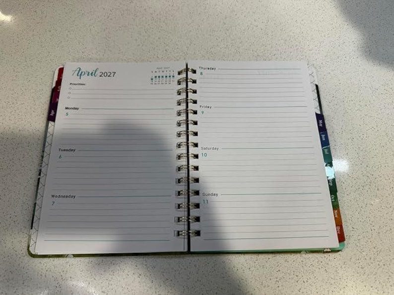 May include: An open spiral-bound planner showing the week of April 5 to April 11, 2027. The left page features a priorities list and days Monday through Wednesday. The right page lists Thursday through Sunday with lined writing spaces.