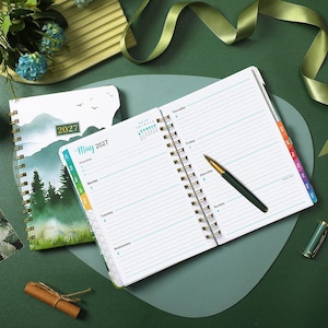 May include: A 2027 spiral-bound planner open to a weekly view for May, featuring lined pages with days of the week printed in blue. A gold and green ballpoint pen rests on the page. The cover displays a mountain and forest landscape illustration.