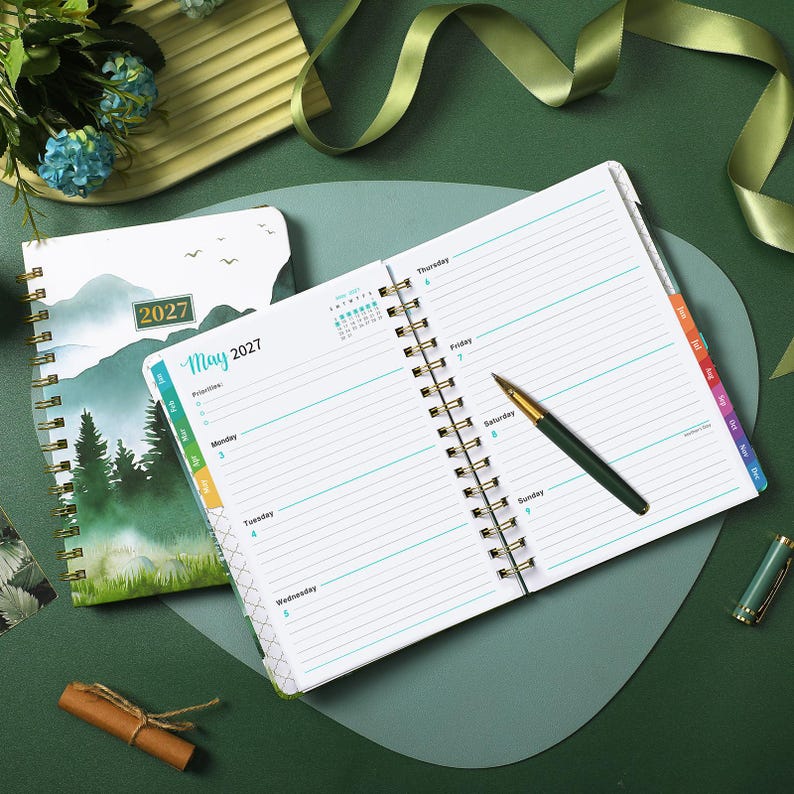 May include: A 2027 spiral-bound planner open to a weekly view for May, featuring lined pages with days of the week printed in blue. A gold and green ballpoint pen rests on the page. The cover displays a mountain and forest landscape illustration.