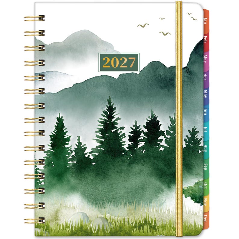 May include: A 2027 spiral-bound planner featuring a watercolor landscape of green pine trees, misty mountains, and birds in flight. The cover includes a gold elastic band closure and colorful monthly tabs along the right edge.