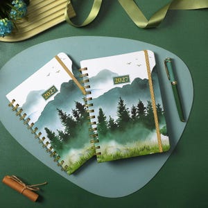 May include: Two spiral-bound planners with a watercolor landscape print of green pine trees, mountains, and birds. Each cover features the text 2027 in a gold-bordered box. A green pen with gold accents lies next to the planners on a green surface.