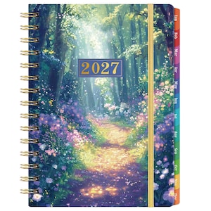 Puede incluir: Una agenda de espiral para 2027 con una escena de bosque, un camino iluminado por el sol, flores silvestres moradas y amarillas y árboles altos. La cubierta incluye un cierre elástico dorado y una etiqueta rectangular con el año 2027 en oro.
