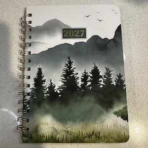 May include: A spiral-bound planner featuring a watercolor-style landscape illustration of dark green pine trees, misty mountains, and birds in flight against a white sky, with the year 2027 printed in a rectangular box on the front cover.
