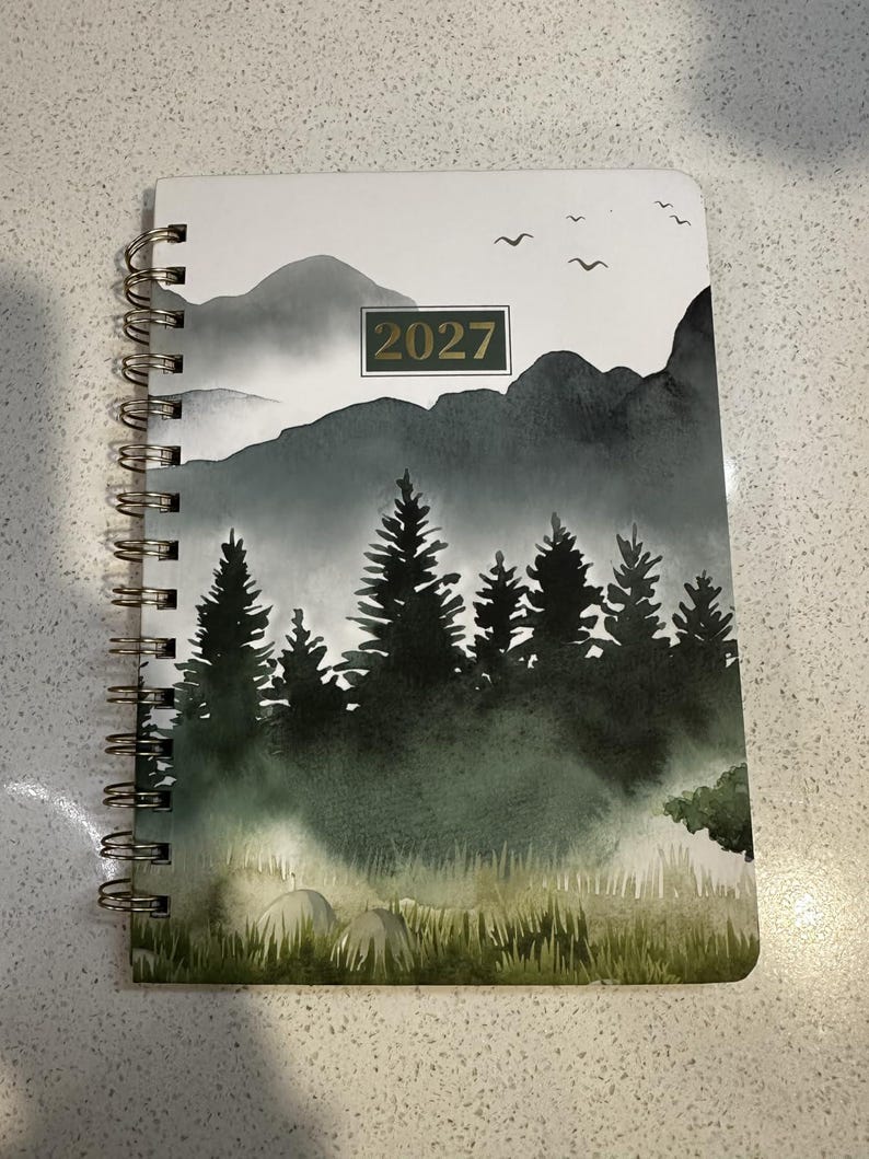 May include: A spiral-bound planner featuring a watercolor-style landscape illustration of dark green pine trees, misty mountains, and birds in flight against a white sky, with the year 2027 printed in a rectangular box on the front cover.