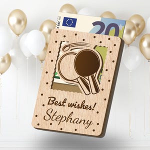 Pu&ograve; includere: Un biglietto di legno con un design ritagliato di due racchette da ping pong. Il biglietto ha il testo "I migliori auguri! Stephany." stampato su di esso.