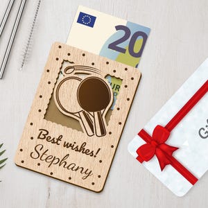 Pu&ograve; includere: Porta biglietti regalo in legno con un design a taglio di due racchette da ping pong. Il porta biglietti ha il testo "I migliori auguri! Stephany" inciso sul davanti. Un biglietto regalo e una banconota da venti euro sono visibili all'interno del porta biglietti.