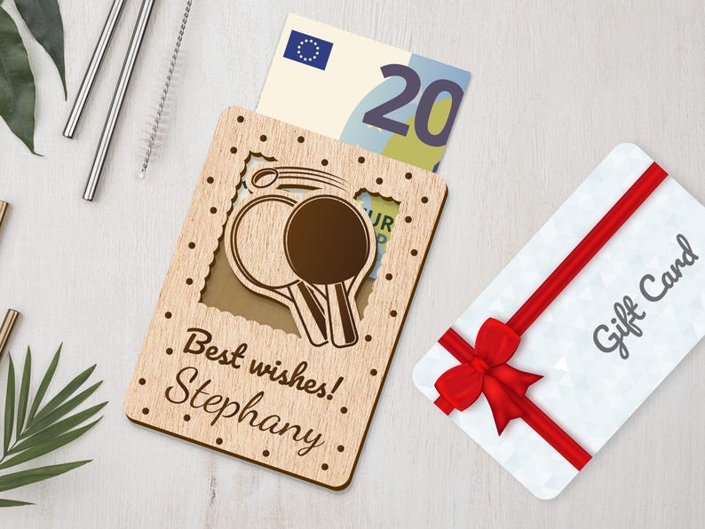 Pu&ograve; includere: Porta biglietti regalo in legno con un design a taglio di due racchette da ping pong. Il porta biglietti ha il testo "I migliori auguri! Stephany" inciso sul davanti. Un biglietto regalo e una banconota da venti euro sono visibili all'interno del porta biglietti.