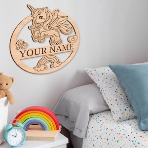 May include: Wooden wall decor featuring a unicorn with a rainbow and clouds. The text "YOUR NAME" is included in the design.