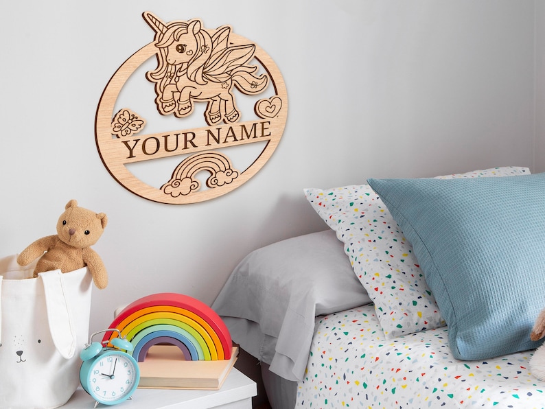 May include: Wooden wall decor featuring a unicorn with a rainbow and clouds. The text "YOUR NAME" is included in the design.