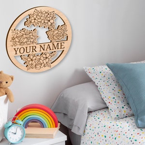 May include: A wooden round sign with a floral design and the text "YOUR NAME" in the center. The sign is hanging on a white wall above a bed with a blue and white patterned pillow.