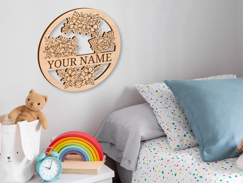 May include: A wooden round sign with a floral design and the text "YOUR NAME" in the center. The sign is hanging on a white wall above a bed with a blue and white patterned pillow.