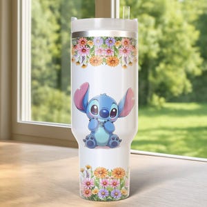Pu&ograve; includere: Un bicchiere bianco con un'immagine a cartoni animati di Stitch, decorato con motivi floreali. Il bicchiere ha un coperchio bianco e una cannuccia trasparente. Il design floreale comprende fiori rosa, arancioni e gialli. Lo sfondo &egrave; una scena esterna verde sfocata.