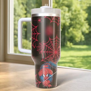 May include: A black and red Spider-Man themed tumbler with a white handle and lid. The tumbler features a red spiderweb design and a graphic of Spider-Man. The tumbler is designed to keep drinks cold or hot.