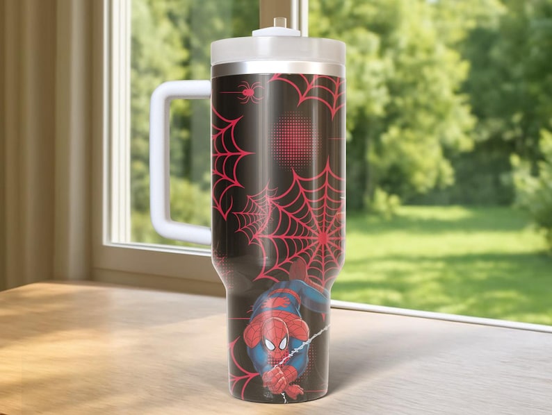 May include: A black and red Spider-Man themed tumbler with a white handle and lid. The tumbler features a red spiderweb design and a graphic of Spider-Man. The tumbler is designed to keep drinks cold or hot.