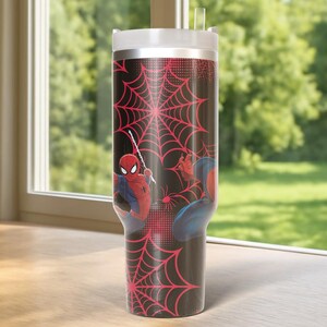 May include: A black tumbler with Spider-Man graphics and a red spiderweb pattern. The tumbler has a white lid and a clear straw. The background shows a window with a view of green trees.