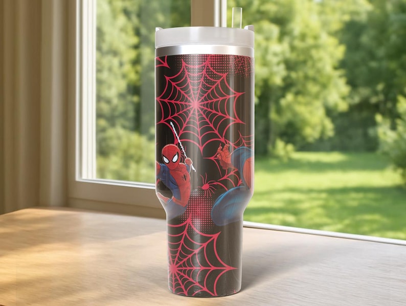 May include: A black tumbler with Spider-Man graphics and a red spiderweb pattern. The tumbler has a white lid and a clear straw. The background shows a window with a view of green trees.