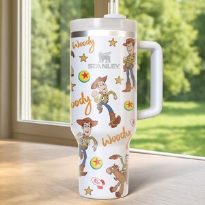 May include: A white Stanley tumbler featuring a Woody design from Toy Story. The tumbler has a white handle and lid, with illustrations of Woody, stars, and the word "Woody" in a playful font. The tumbler is approximately 10 inches tall.