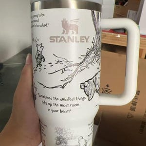 Puede incluir: Vaso Stanley blanco con un asa grande, con ilustraciones de personajes de Winnie the Pooh y citas. El vaso tiene un borde plateado y una tapa blanca. El texto "STANLEY" es visible. La cita "Sometimes the smallest things take up the most room in your heart" tambi&eacute;n es visible.