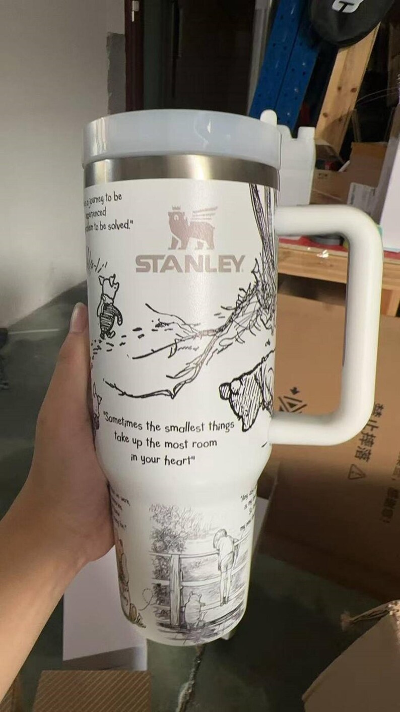 Puede incluir: Vaso Stanley blanco con un asa grande, con ilustraciones de personajes de Winnie the Pooh y citas. El vaso tiene un borde plateado y una tapa blanca. El texto "STANLEY" es visible. La cita "Sometimes the smallest things take up the most room in your heart" tambi&eacute;n es visible.