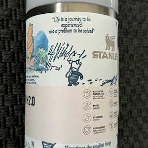 Puede incluir: Un termo de viaje Stanley H2.0 blanco y plateado con ilustraciones y citas de Winnie the Pooh. El termo incluye el texto "Life is a journey to be experienced not a problem to be solved" y "Sometimes the smallest things take up the most room in your heart."