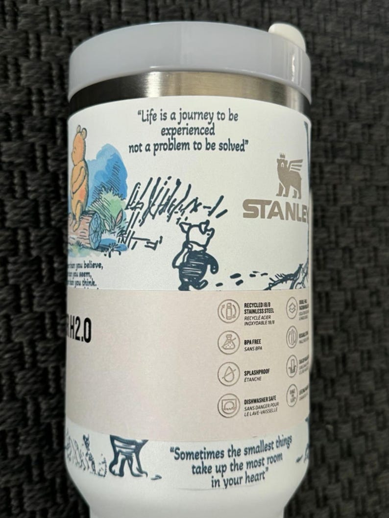 Puede incluir: Un termo de viaje Stanley H2.0 blanco y plateado con ilustraciones y citas de Winnie the Pooh. El termo incluye el texto "Life is a journey to be experienced not a problem to be solved" y "Sometimes the smallest things take up the most room in your heart."