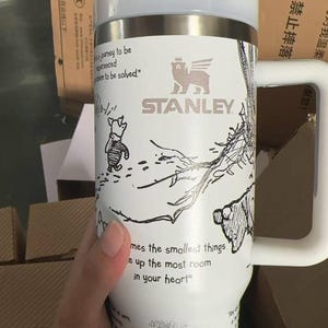 Puede incluir: Vaso Stanley blanco con asa, con un dise&ntilde;o de Winnie the Pooh. El vaso tiene una tapa blanca y una banda de acero inoxidable. El texto incluye "Stanley" y una cita: "mes the smallest things e up the most room in your heart."