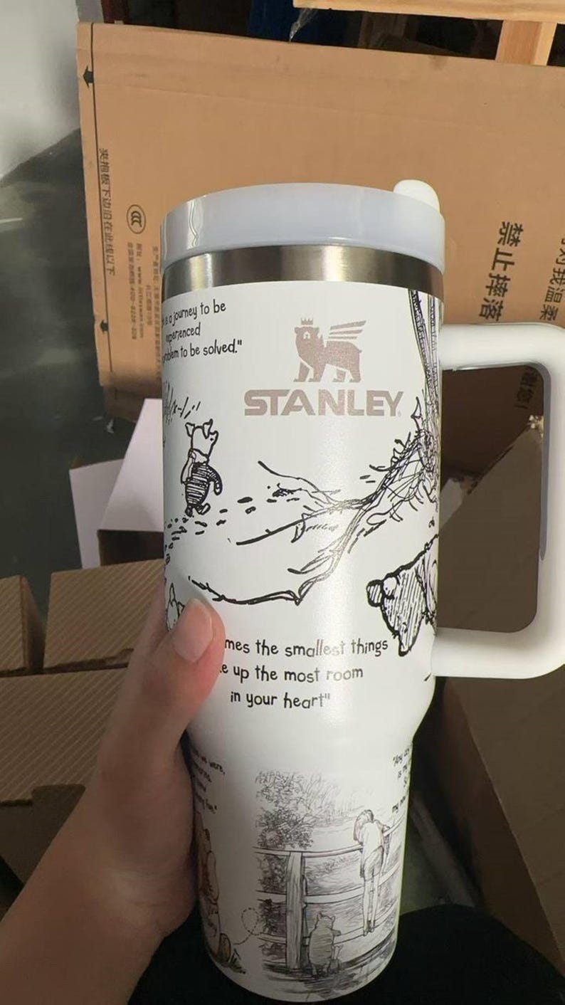 Puede incluir: Vaso Stanley blanco con asa, con un dise&ntilde;o de Winnie the Pooh. El vaso tiene una tapa blanca y una banda de acero inoxidable. El texto incluye "Stanley" y una cita: "mes the smallest things e up the most room in your heart."