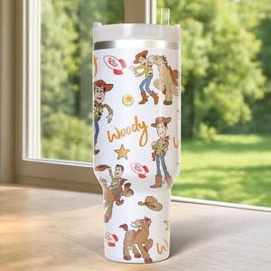 May include: A white tumbler featuring cartoon images of Woody from Toy Story. The tumbler has a silver rim and a white lid with a clear straw. The design includes Woody, a horse, and other Toy Story elements.