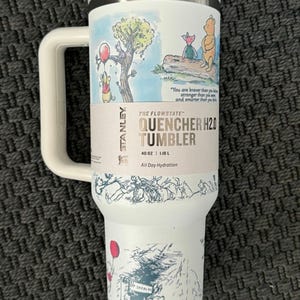 Puede incluir: Vaso Stanley Flowstate Quencher H2.0 blanco con asa, con ilustraciones de Winnie the Pooh. El vaso tiene una tapa y asa grises, y el texto "QUENCHER H2.0 TUMBLER" est&aacute; impreso en el lateral. Capacidad: 1,18 L.