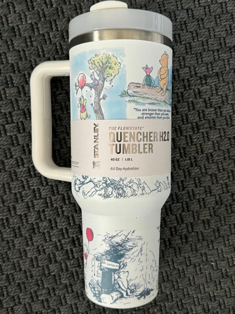 Puede incluir: Vaso Stanley Flowstate Quencher H2.0 blanco con asa, con ilustraciones de Winnie the Pooh. El vaso tiene una tapa y asa grises, y el texto "QUENCHER H2.0 TUMBLER" est&aacute; impreso en el lateral. Capacidad: 1,18 L.