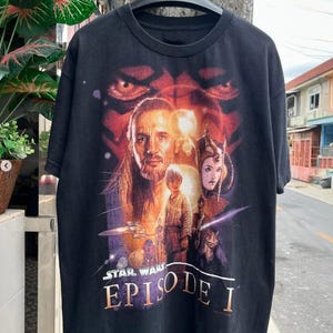 May include: Black t-shirt featuring a Star Wars Episode I graphic. The design includes characters and the movie title in orange and white. The shirt is hanging on a black hanger.