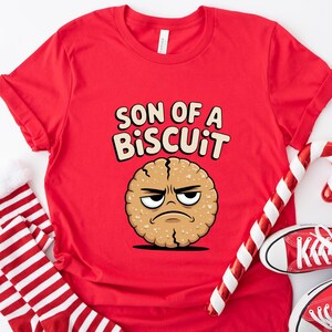 May include: A red t-shirt with the text "SON OF A BISCUIT" and a cartoon biscuit character. The image also includes red and white striped socks, a candy cane, and red sneakers.