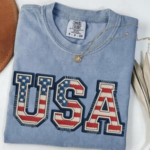 May include: A light blue Comfort Colors t-shirt with the word "USA" in a patriotic design. The letters are outlined in navy blue with a red, white, and blue flag pattern. A gold necklace and a brown hat are also visible.